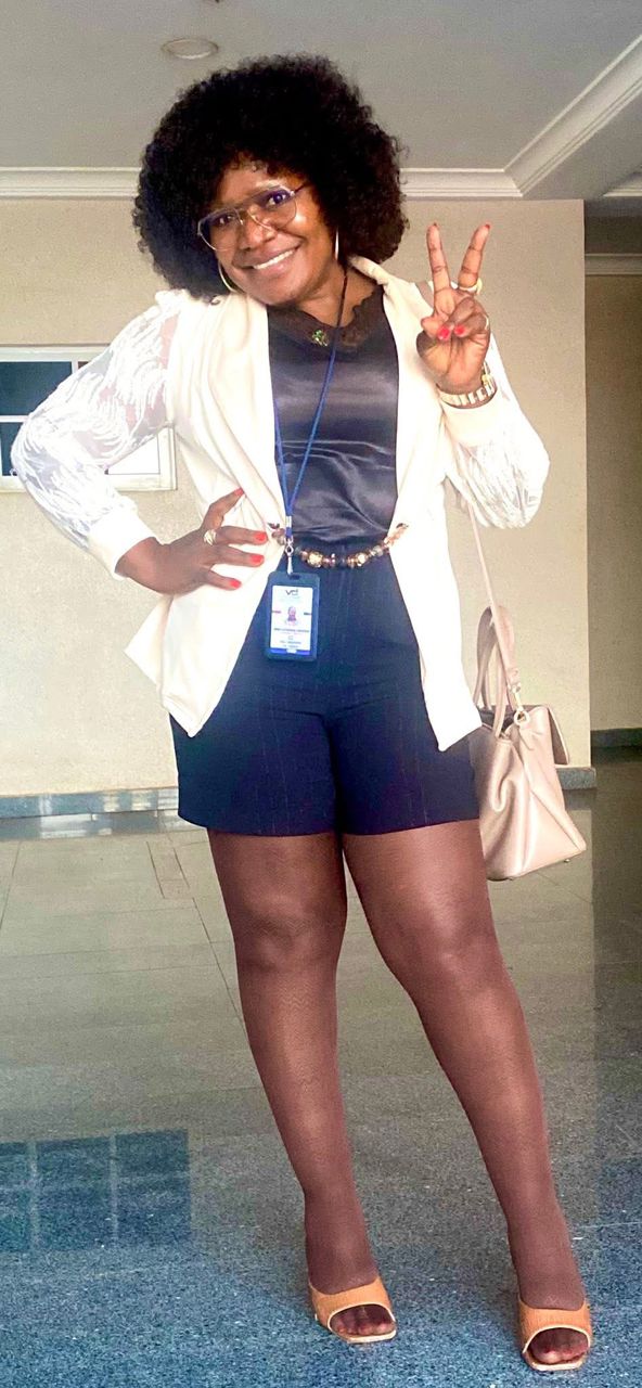 An image of Cathrine Chi Edeh, smiling indoors, posing with one hand on her hip and the other hand making a peace sign. She is wearing glasses, a white blazer with lace sleeves, a black blouse, black shorts, sheer tights, and open-toe tan heels. She has a lanyard with an ID badge around her neck and a light beige handbag hanging from her shoulder. She has curly hair and is standing on a shiny tiled floor, with a light-colored wall in the background.