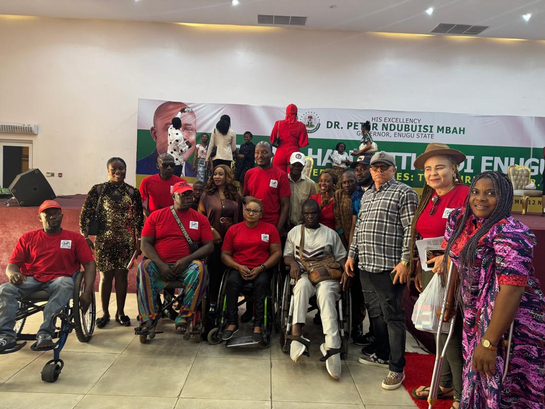 An image of a group of people, including men and women, gathered indoors for a group photo. Several individuals are seated in wheelchairs, and a few others are using crutches, indicating the presence of persons with disabilities. Some of the people are wearing matching red T-shirts and caps, while others are in casual or formal attire. The background features a banner that reads "His Excellency Dr. Peter Ndubuisi Mbah Governor, Enugu State" and there’s an image of a man’s face on the banner. The atmosphere appears to be that of an event or gathering, possibly organized in recognition or support of people with disabilities. The setting is a large hall with a stage and sound equipment visible.