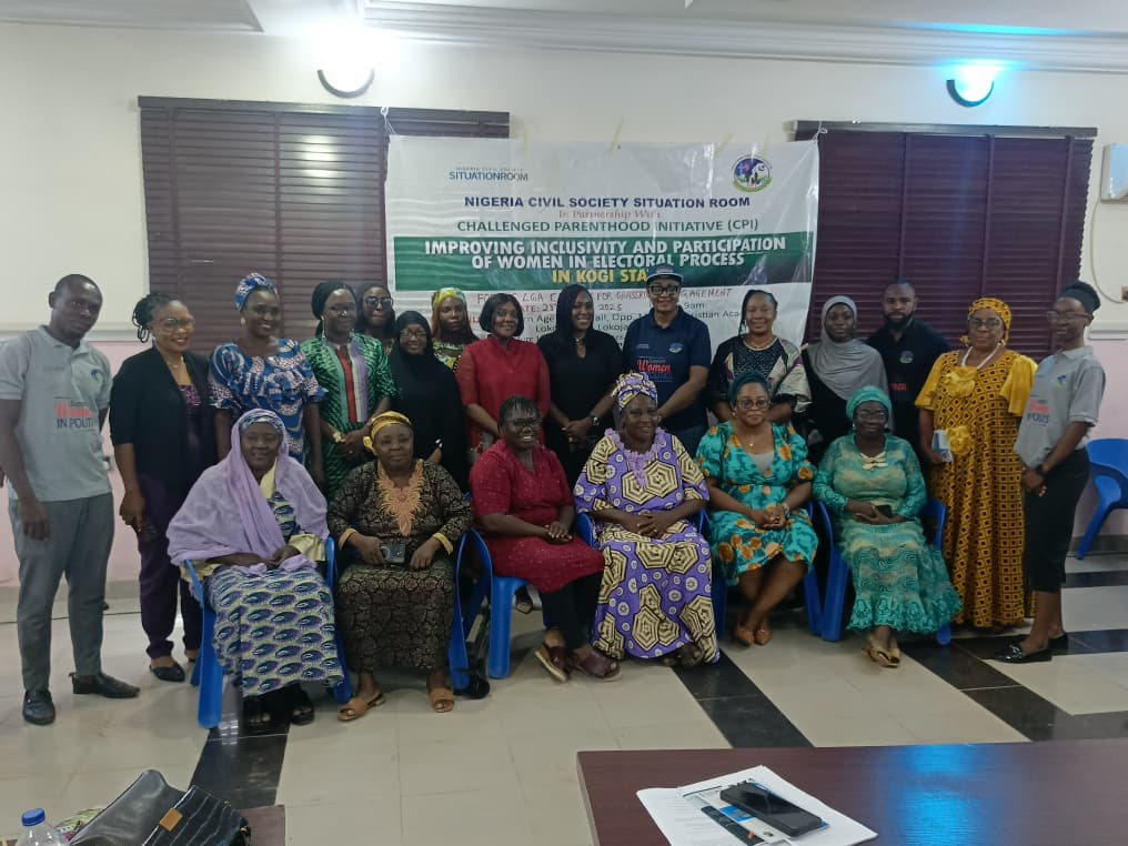 An image of a group of men and women posing together indoors. There are 21 people, with seven women sitting in the front row and the rest standing behind them. Most are dressed in traditional African clothing, while a few wear modern attire. Behind them hangs a large banner that reads:

"NIGERIA CIVIL SOCIETY SITUATION ROOM
CHALLENGED PARENTHOOD INITIATIVE (CPI)
IMPROVING INCLUSIVITY AND PARTICIPATION
OF WOMEN IN ELECTORAL PROCESS
IN KOGI STATE"

The room has blue plastic chairs and brown window blinds. There is a table in the foreground with some documents and items on it. The atmosphere suggests a formal group meeting or workshop focusing on women's participation in electoral processes.