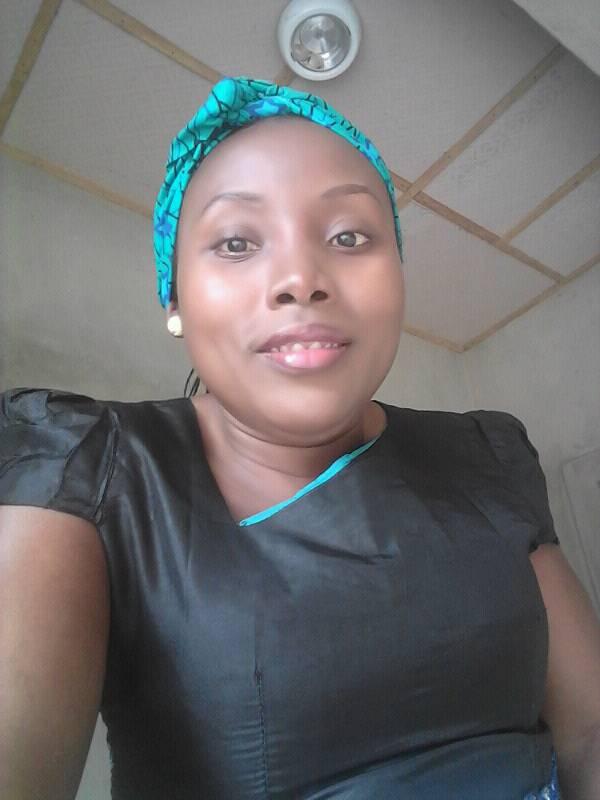 An image of Mary in a black dress with a turquoise and black patterned headscarf. She is smiling slightly and looking straight at the camera. The background shows a ceiling with a light fixture and cream-colored walls. She is wearing pearl stud earrings. The atmosphere of the photo is bright and casual.